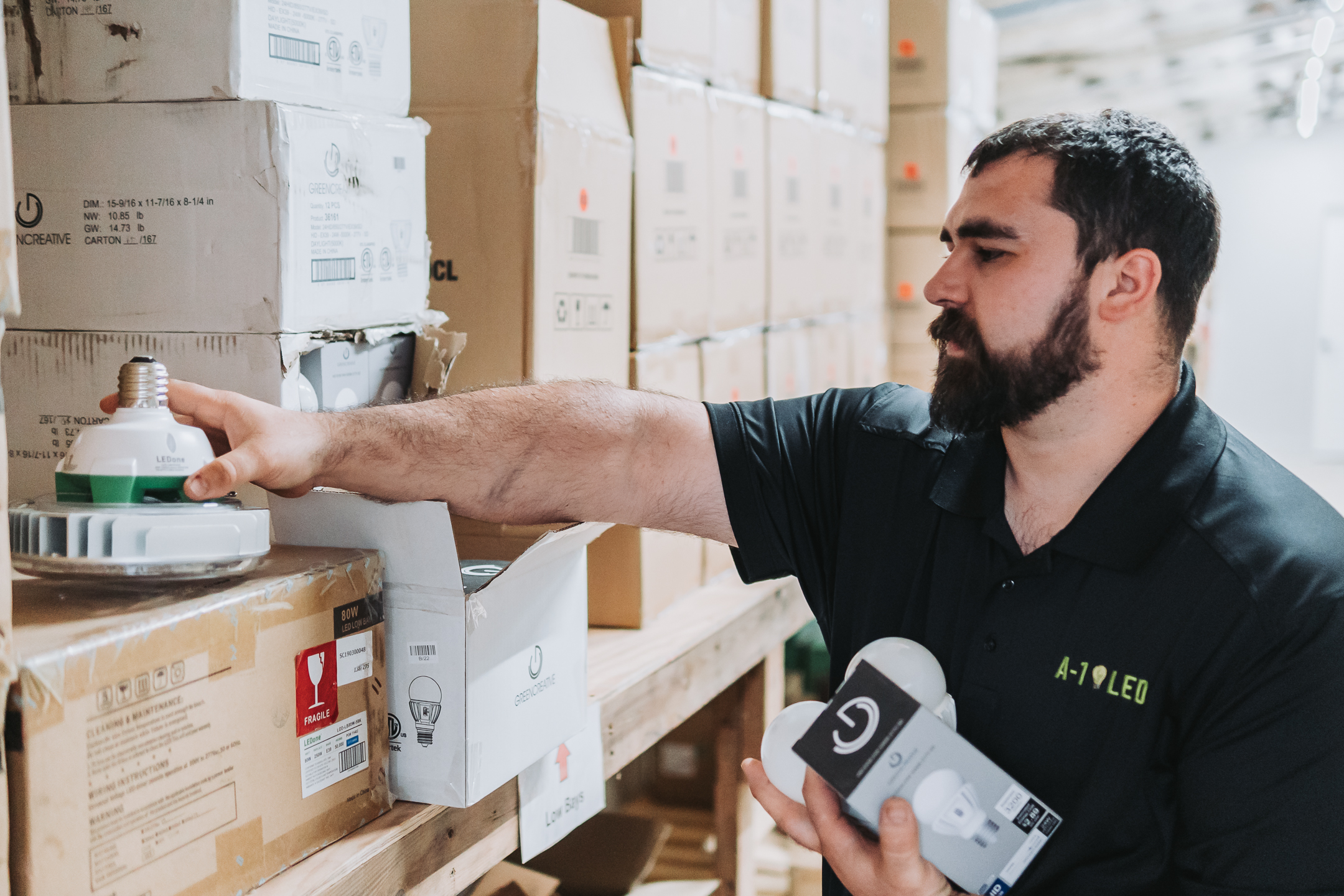A man with a beard wearing a black shirt selects an LED light bulb from a shelf filled with cardboard boxes in a warehouse. He holds two boxes in one hand while reaching for another bulb.