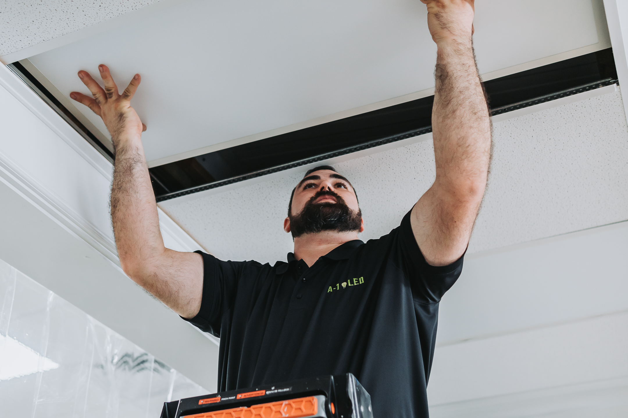 A man with a beard stands on a stepladder, installing or adjusting a ceiling tile in an indoor space. He wears a black shirt with green text and reaches up with both hands to fit the panel.