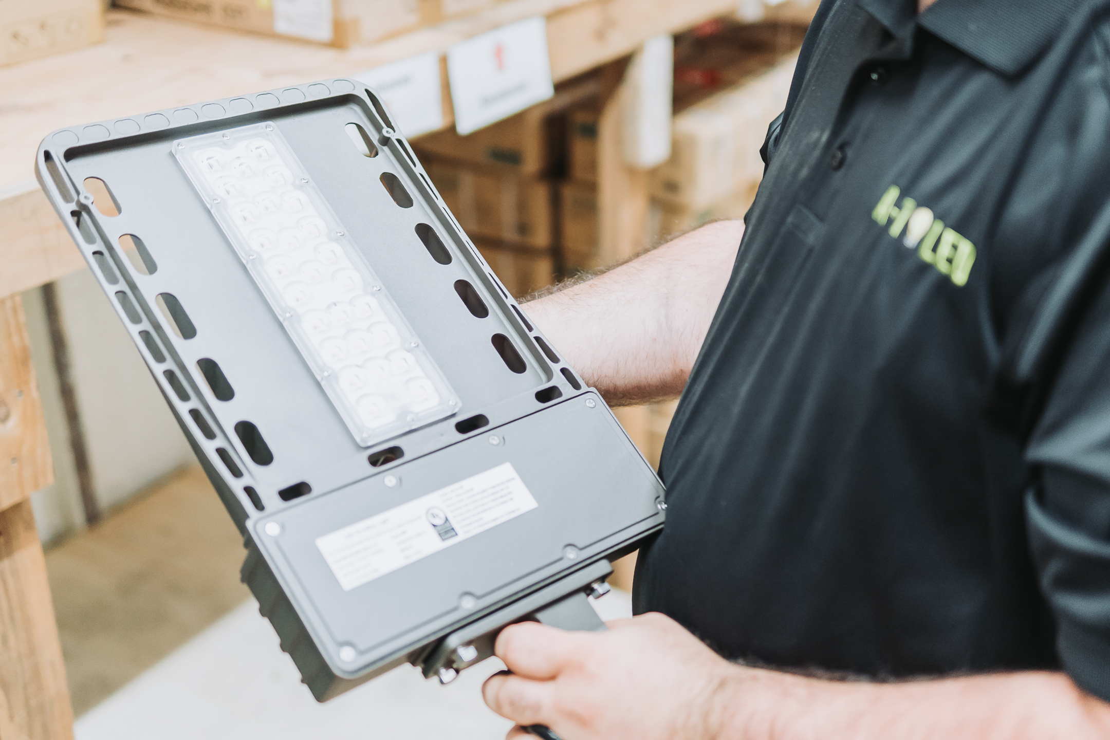 A person in a black polo shirt holds an LED light fixture in a warehouse setting with shelves and boxes visible in the background.