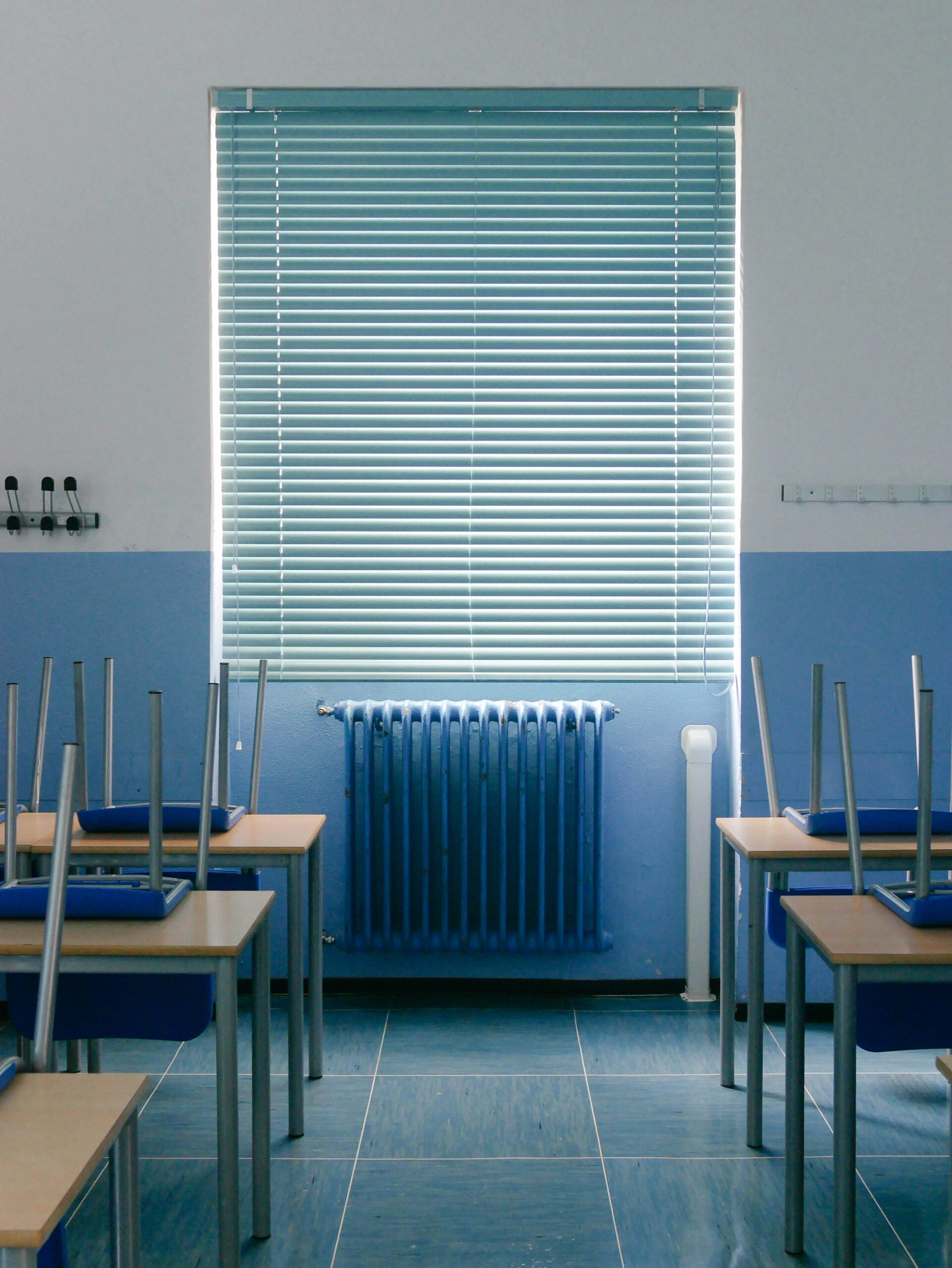 Empty classroom with desks and chairs neatly arranged, chairs upside down on desks, blue radiator, window blinds closed. Great example for LED lighting tips on energy efficiency in schools.