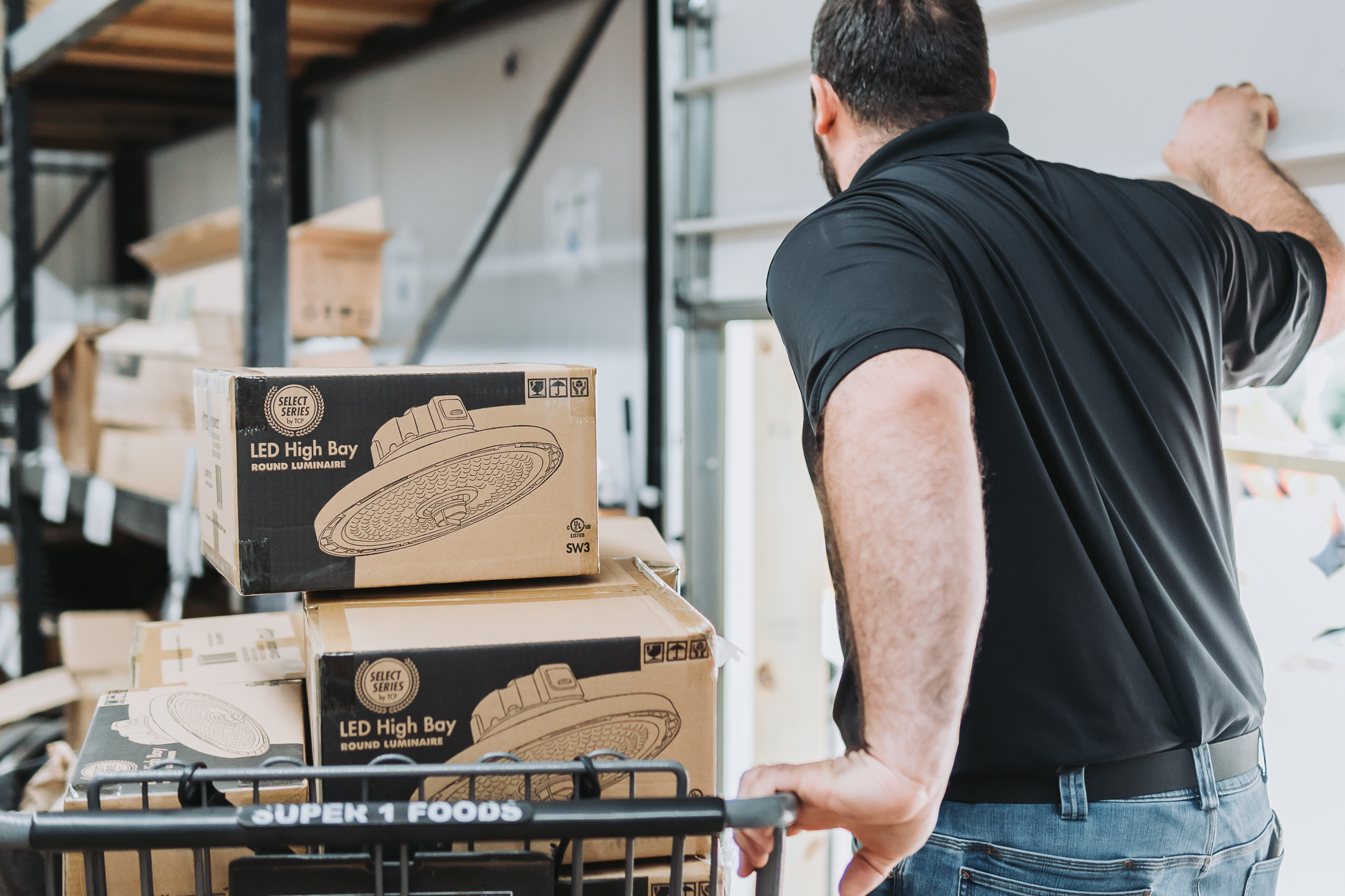 A man in a black shirt pushes a cart with boxed LED high bay lights in a warehouse, with shelves and packages in the background.