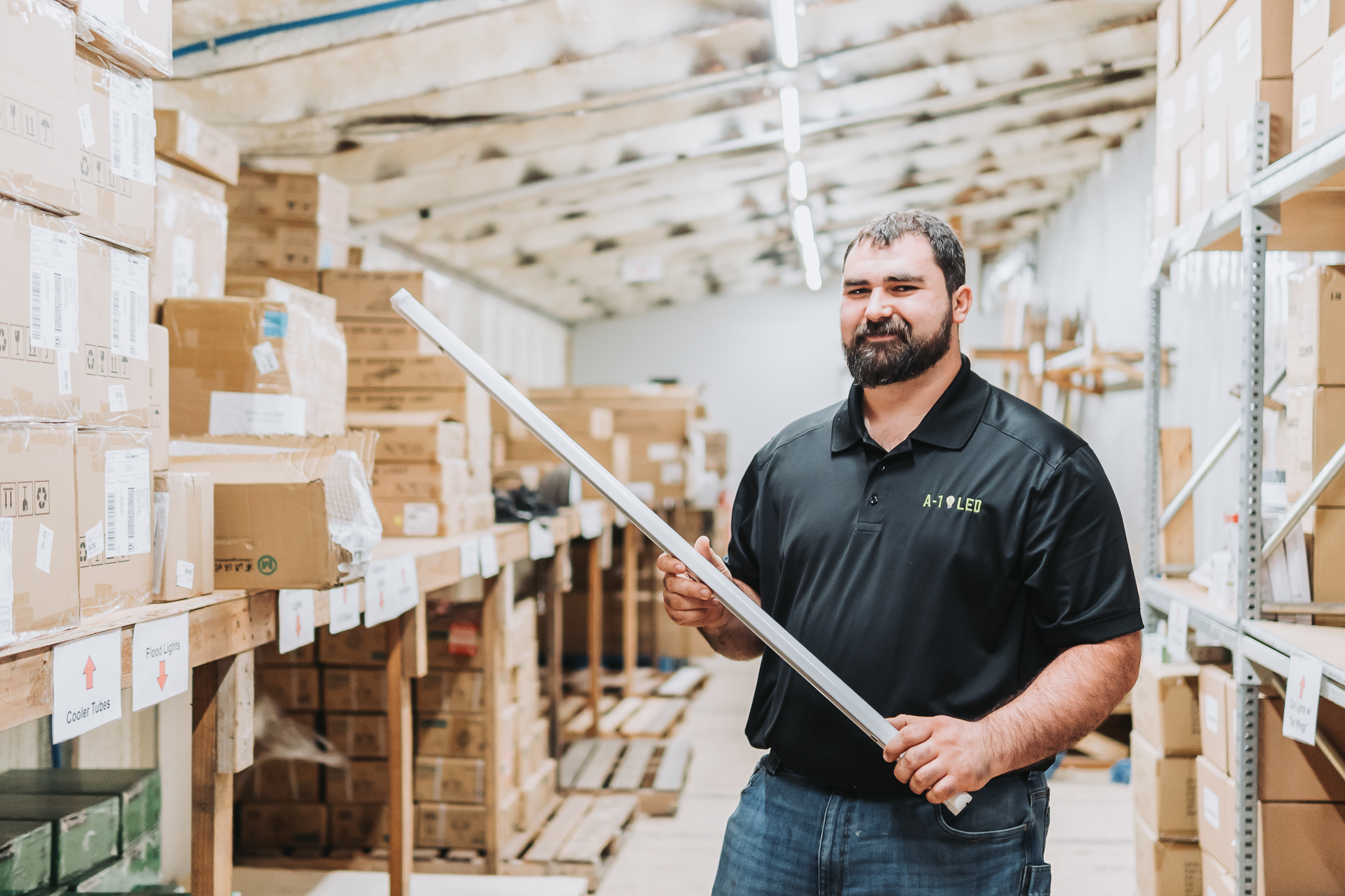 A man in a black polo shirt stands in a warehouse filled with cardboard boxes, holding a long metal rod and smiling at the camera. Halloween lighting casts playful shadows as shelves with labeled boxes line both sides of the aisle.