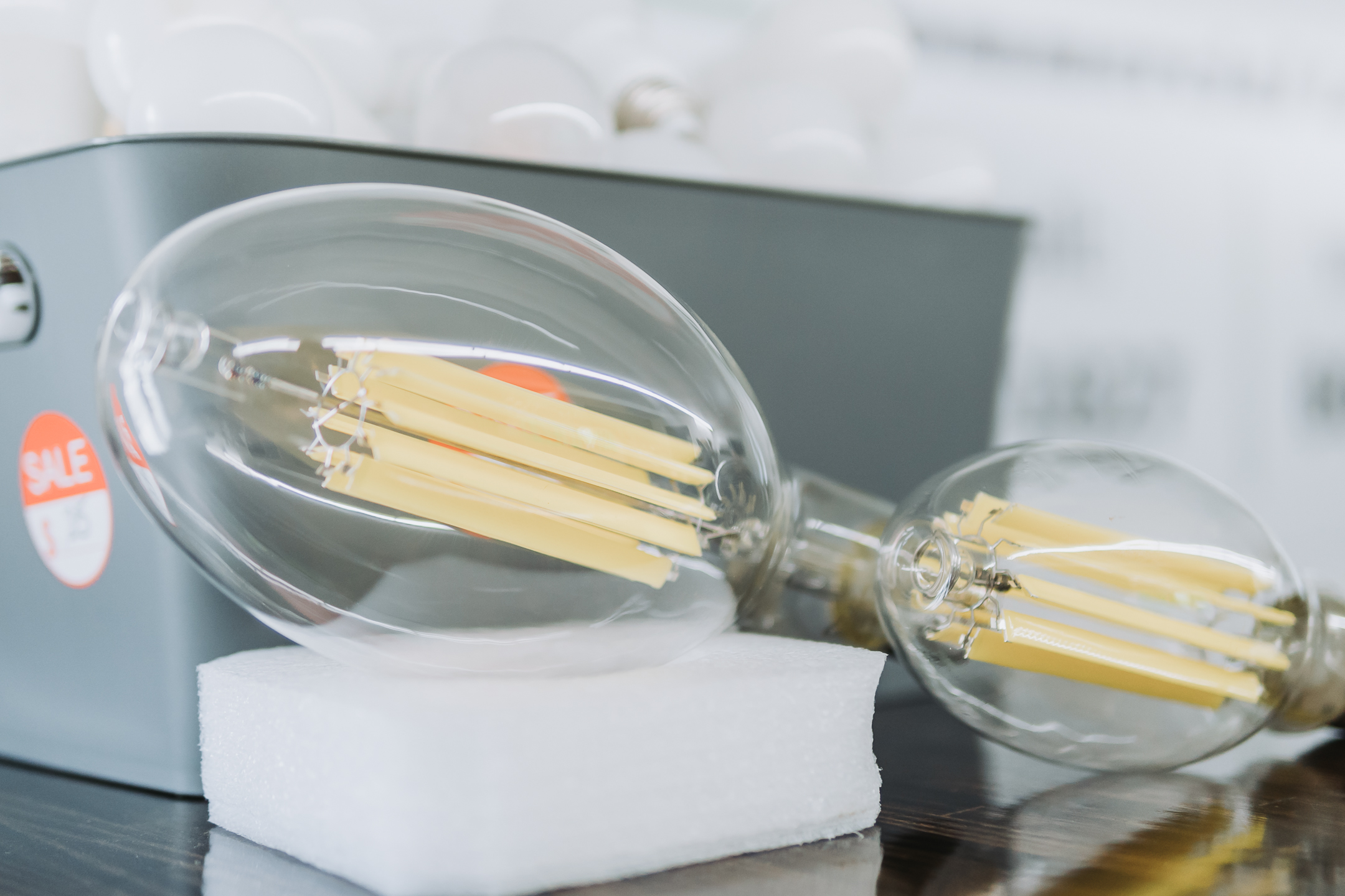 Close-up of two clear LED filament light bulbs, one on white foam and the other leaning against a gray SALE-marked box. Perfect for brightening up your space as daylight savings time approaches. More bulbs are visible in the background.