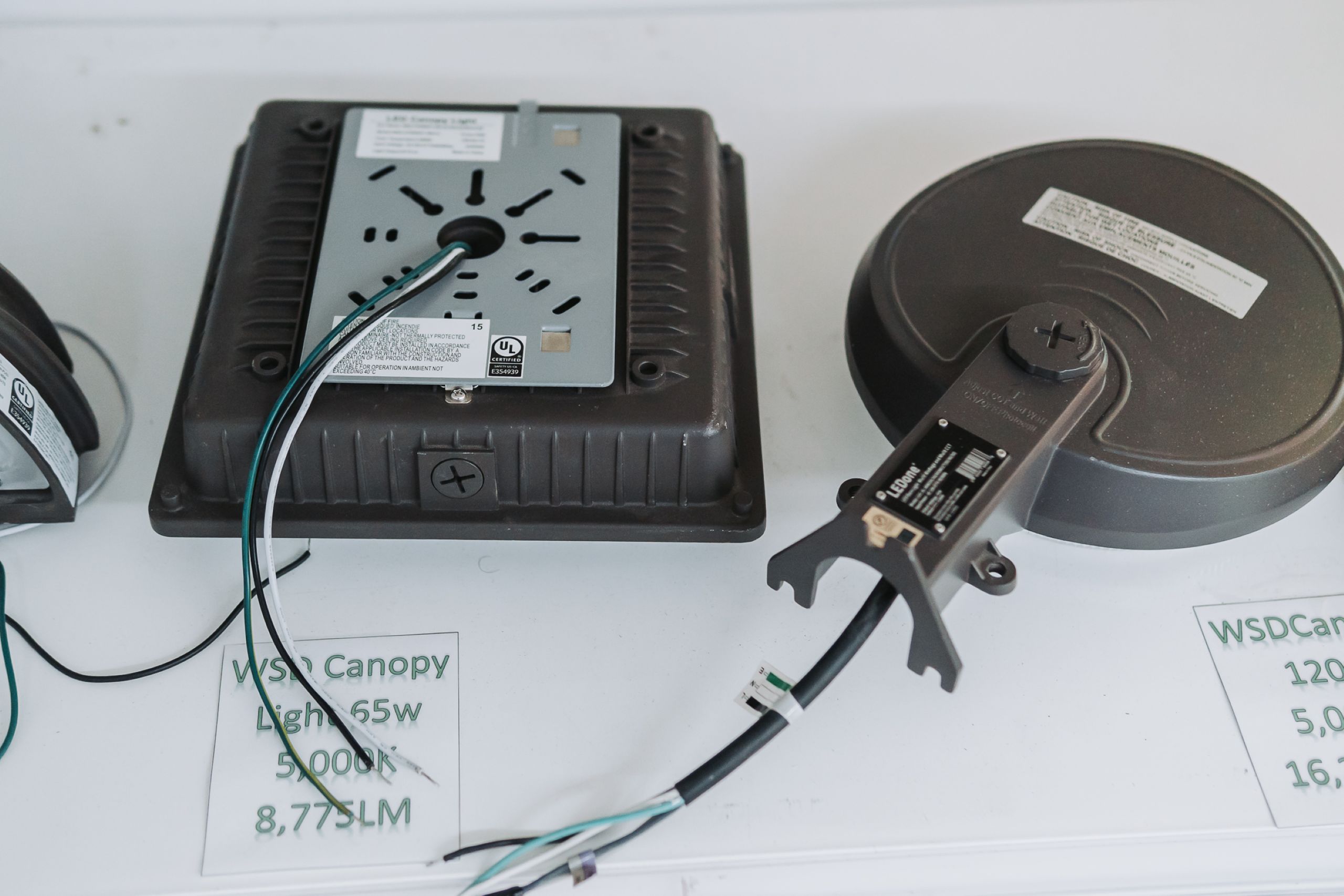 Close-up of two industrial LED canopy light fixtures placed on a white surface, with exposed wiring and information labels visible next to printed product specifications.