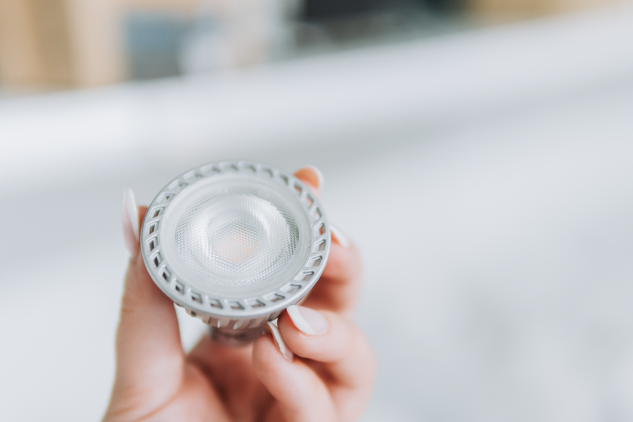 A close-up of a hand holding a small, round LED spotlight bulb with a textured glass cover and silver rim, highlighting how LED lighting can improve safety during winter against a blurred white background.