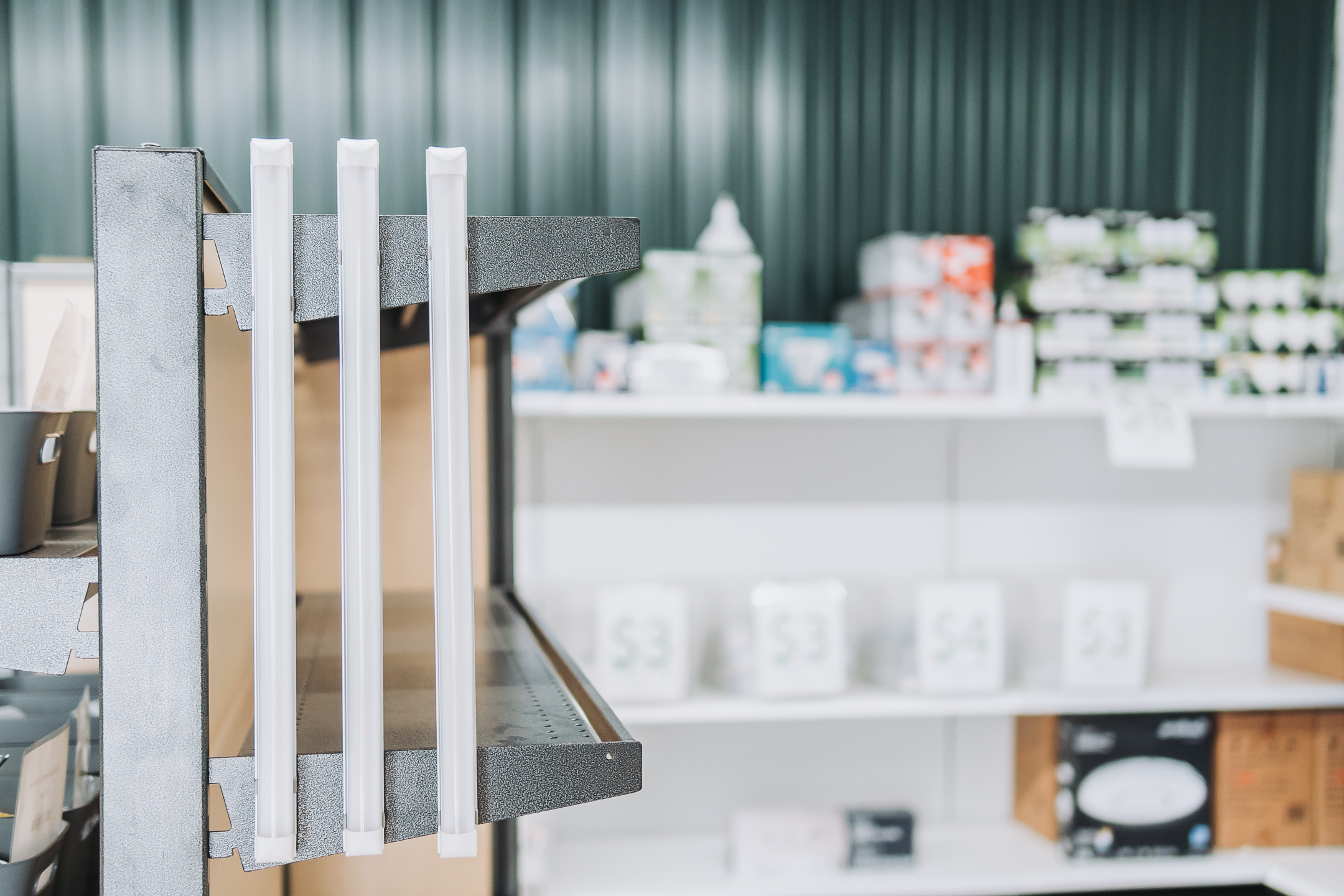 A close-up of an empty metal shelf with three white dividers in a store, illuminated by commercial-grade LED lighting; blurred shelves with various boxes and products are visible in the background.