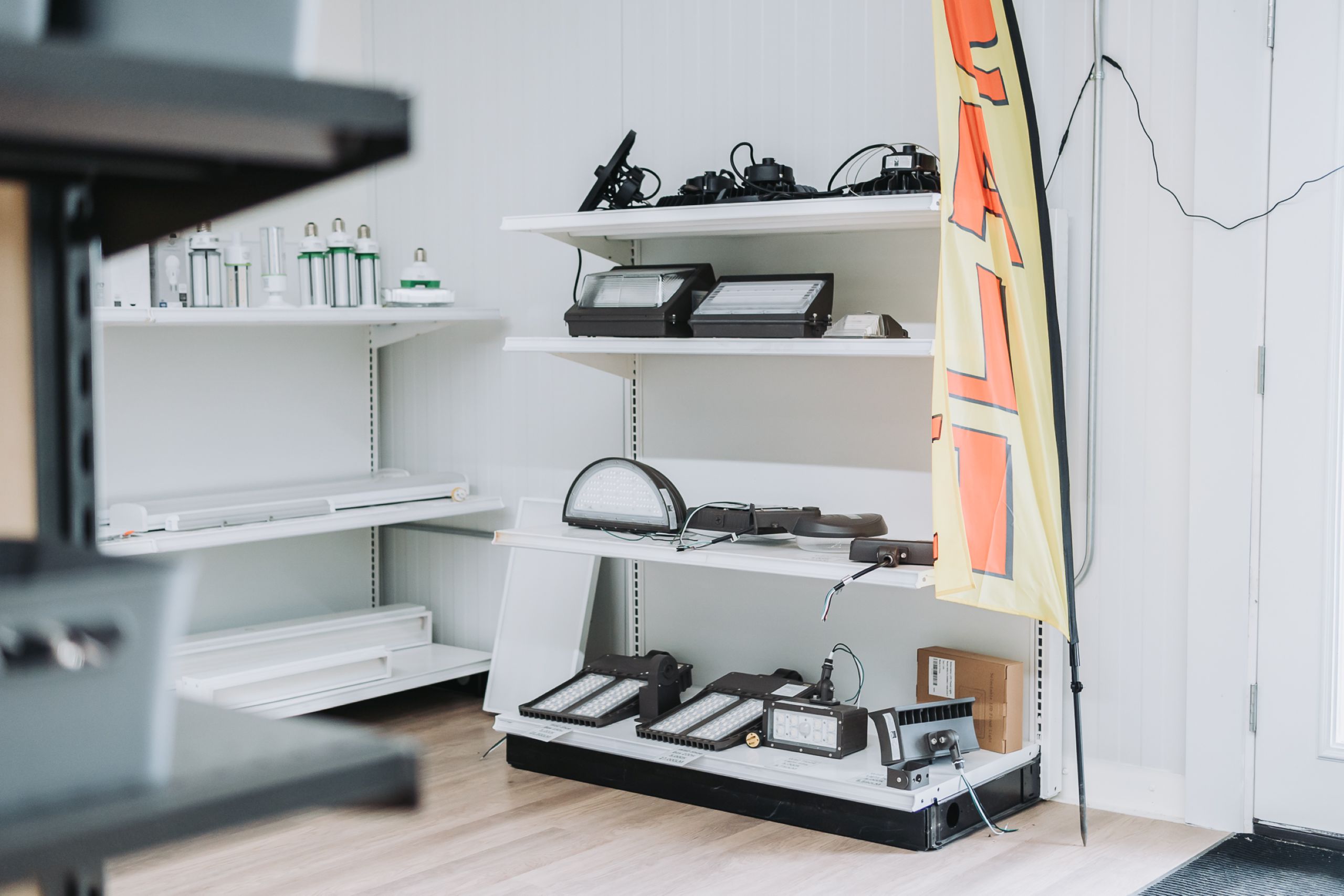 Shelves displaying various LED lights and lighting equipment inside a store, with a yellow and red flag leaning against the wall. As your trusted Lighting Partner, we make finding quality solutions and electrical cords easy.