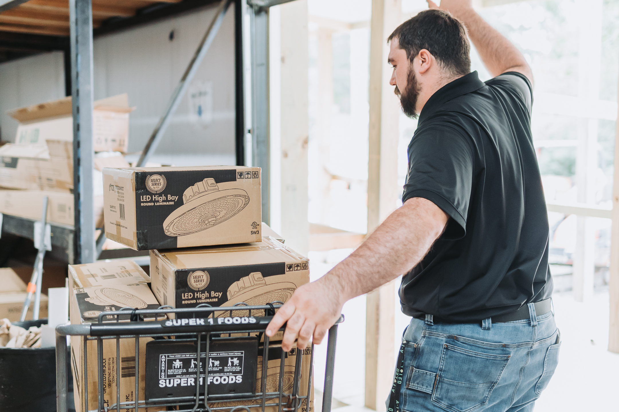 A man in a black shirt pushes a cart labeled Super 1 Foods loaded with several boxes of LED high bay lights and LED flood lights inside a warehouse or storage area, highlighting commercial led flood lights