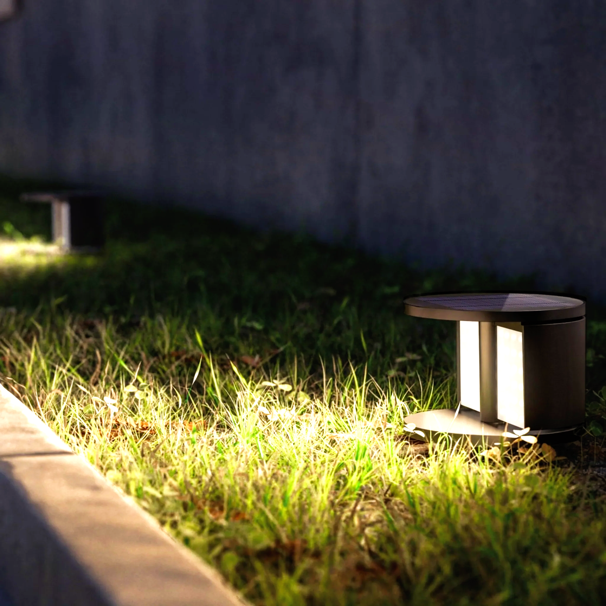 Two small, modern outdoor solar lights, part of stylish LED lighting solutions, illuminate grass along a concrete curb, casting a warm glow in the evening near a gray wall.