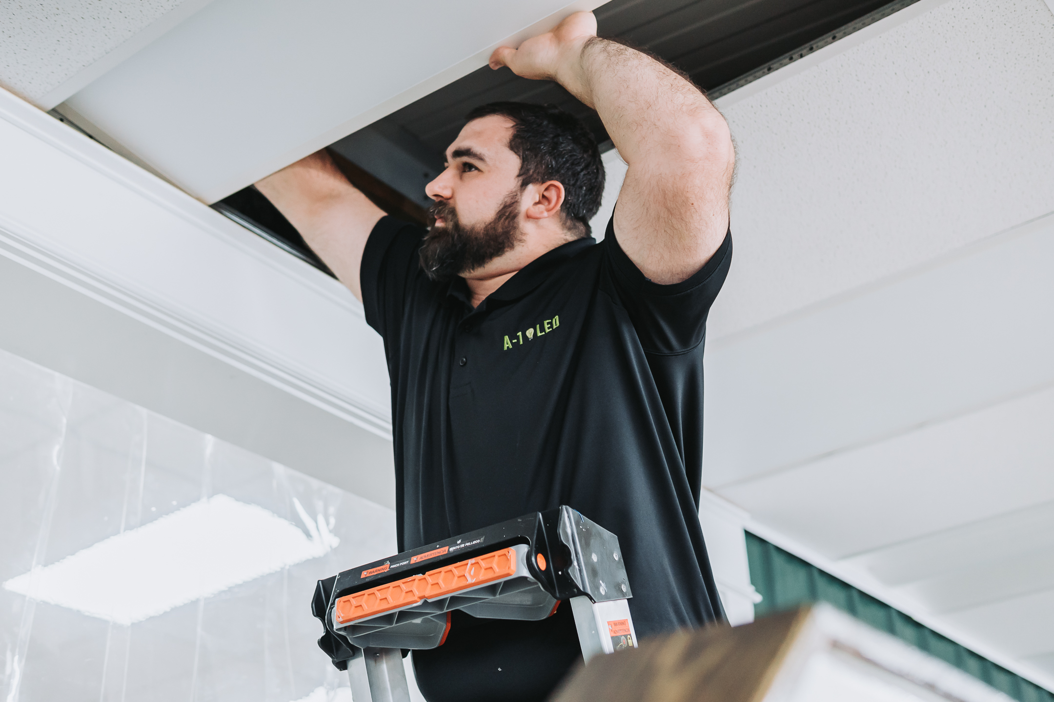 Spring Cleaning for Your Lighting Maintenance Tips for LED Fixtures A man standing on a ladder doing led lighting maintenance. He repairs a ceiling panel and works on the lighting in an indoor space. He is wearing a black polo shirt with A-1 LEX printed on it.