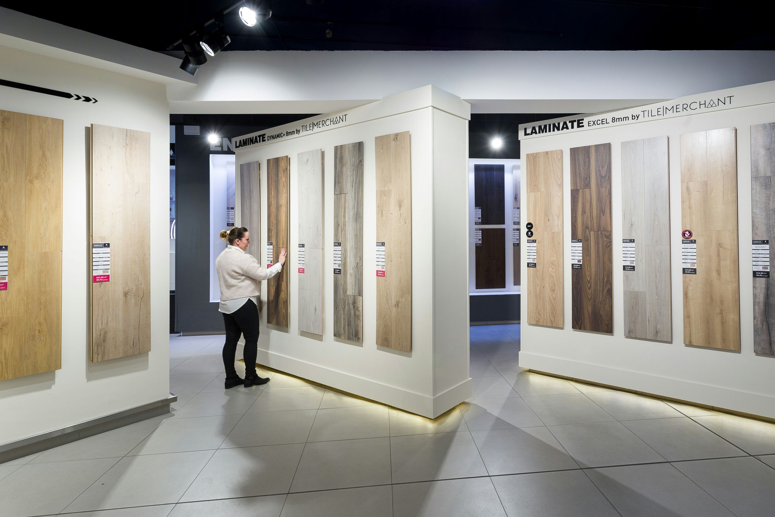 A person stands in a showroom looking at large samples of laminate flooring mounted on white display walls illuminated by bright LED fixtures. The flooring samples vary in color and wood grain patterns.