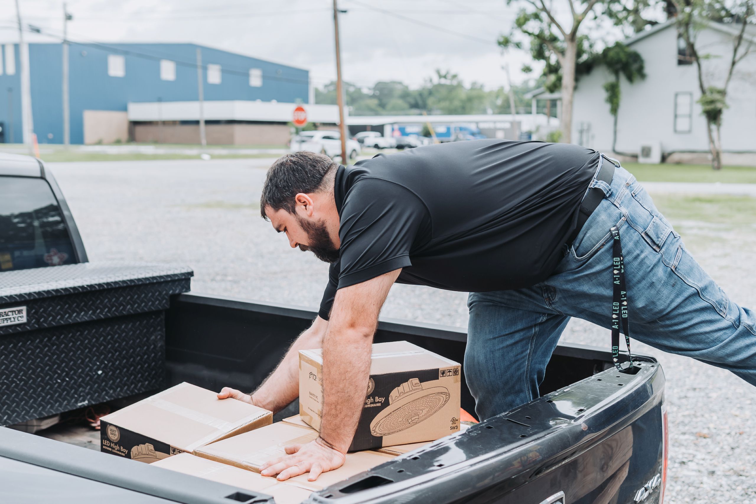 Why May Is Ideal for LED Upgrades Man in black shirt and jeans bends over to load boxes from a truck bed. One box shows LED High Bay Round Luminaire.