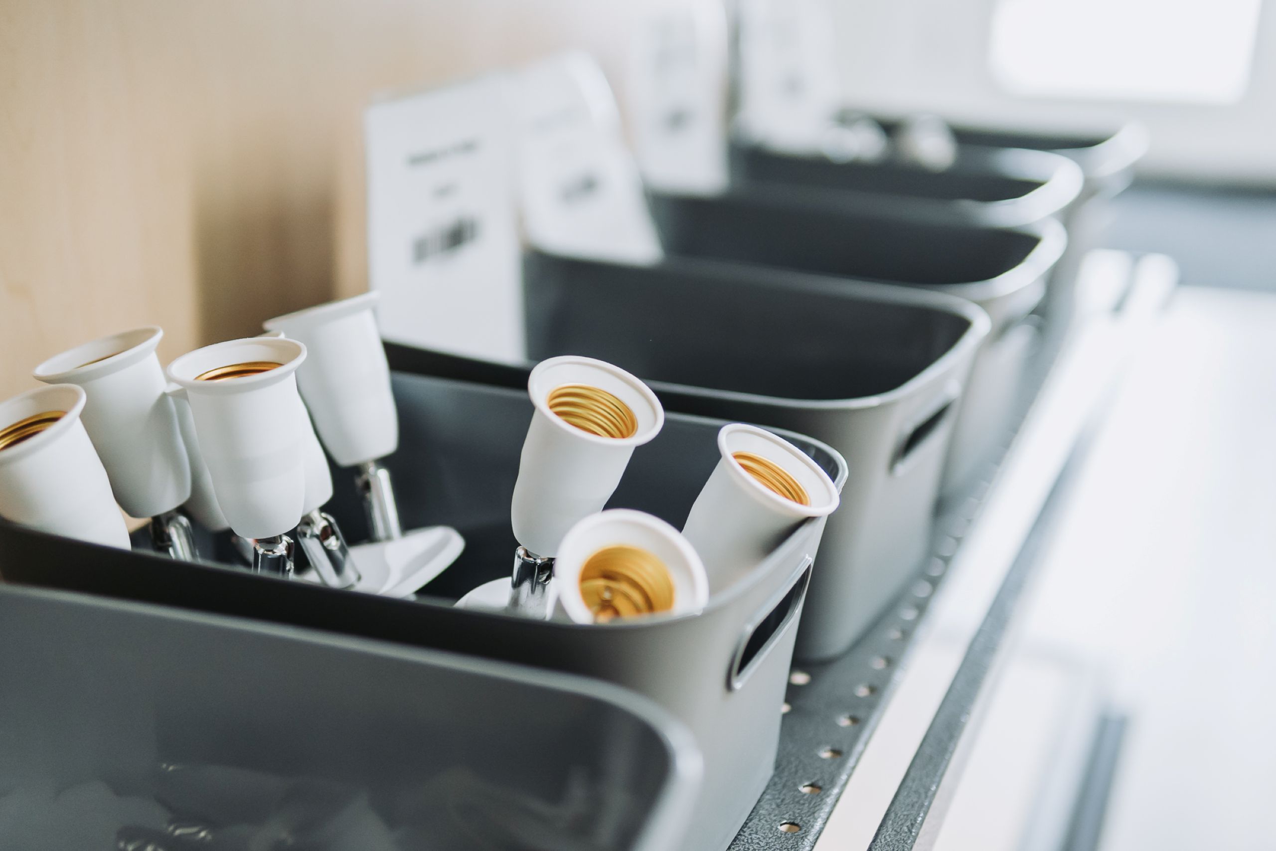 Gray bins on a shelf hold several white light bulb sockets, neatly arranged in rows, with more empty bins visible in the background. The organized scene suggests a focus on energy efficient lighting solutions.